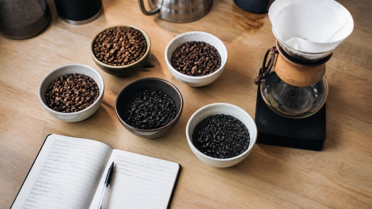 Overhead flat lay of several small bowls of whole coffee beans in different roast shades, a handwritten tasting notebook, and a pour-over cone on a light wood kitchen counter